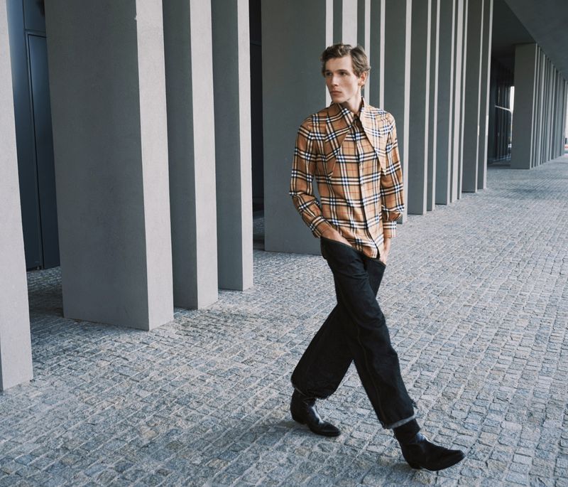 An adult male model wearing a plaid shirt and black trousers is walking on a city street with modern architecture during daytime.