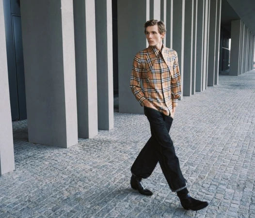 An adult male model wearing a plaid shirt and black trousers is walking on a city street with modern architecture during daytime.