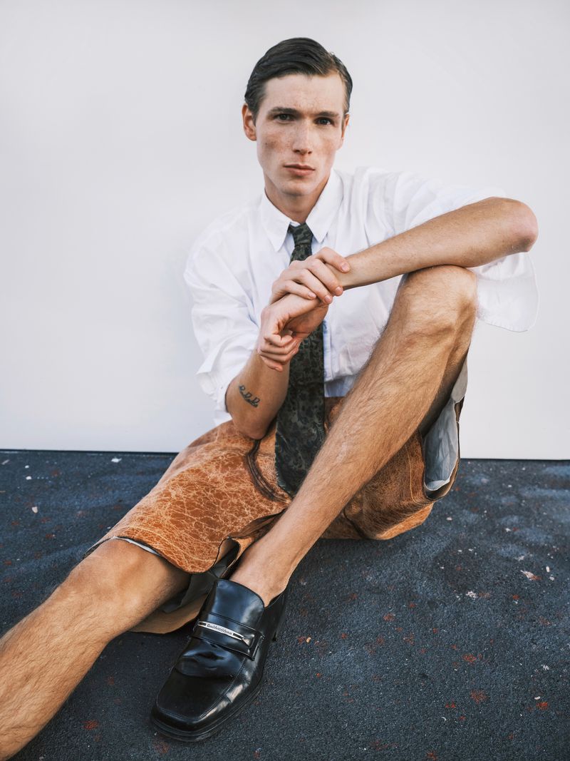 Stylish young adult male sitting with leather lederhosen and dress shirt in a fashion pose against a white wall background.