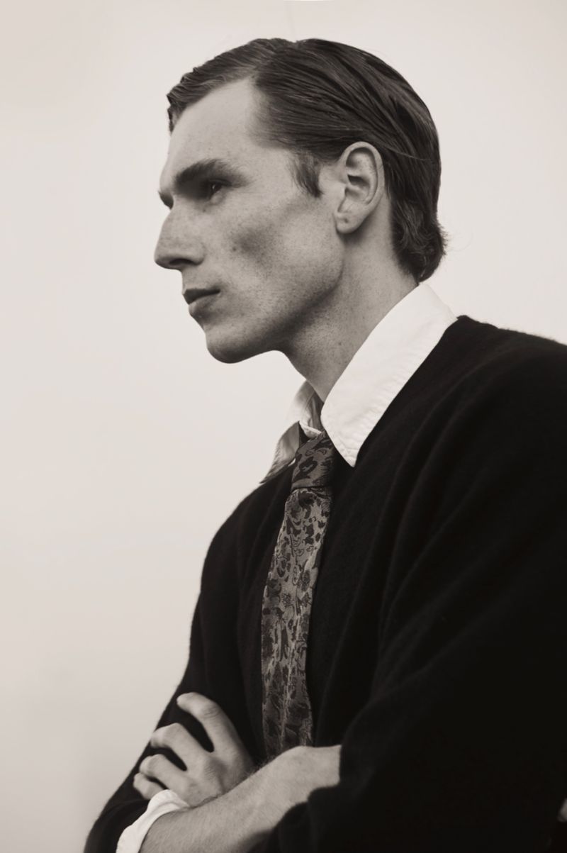 Portrait of a man in a black sweater with folded arms, wearing a patterned tie and white collared shirt against a light background.