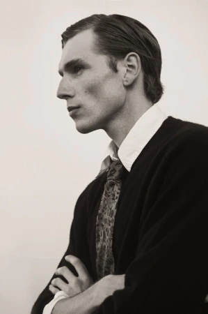 Portrait of a man in a black sweater with folded arms, wearing a patterned tie and white collared shirt against a light background.