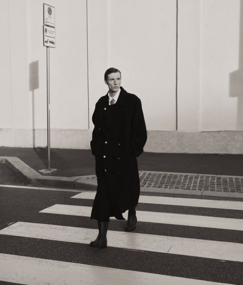 A stylish man in a dark overcoat strides confidently across a crosswalk, set against a minimalist urban backdrop with bright lighting.