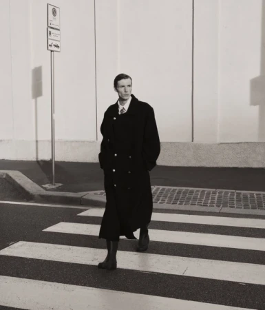 A stylish man in a dark overcoat strides confidently across a crosswalk, set against a minimalist urban backdrop with bright lighting.