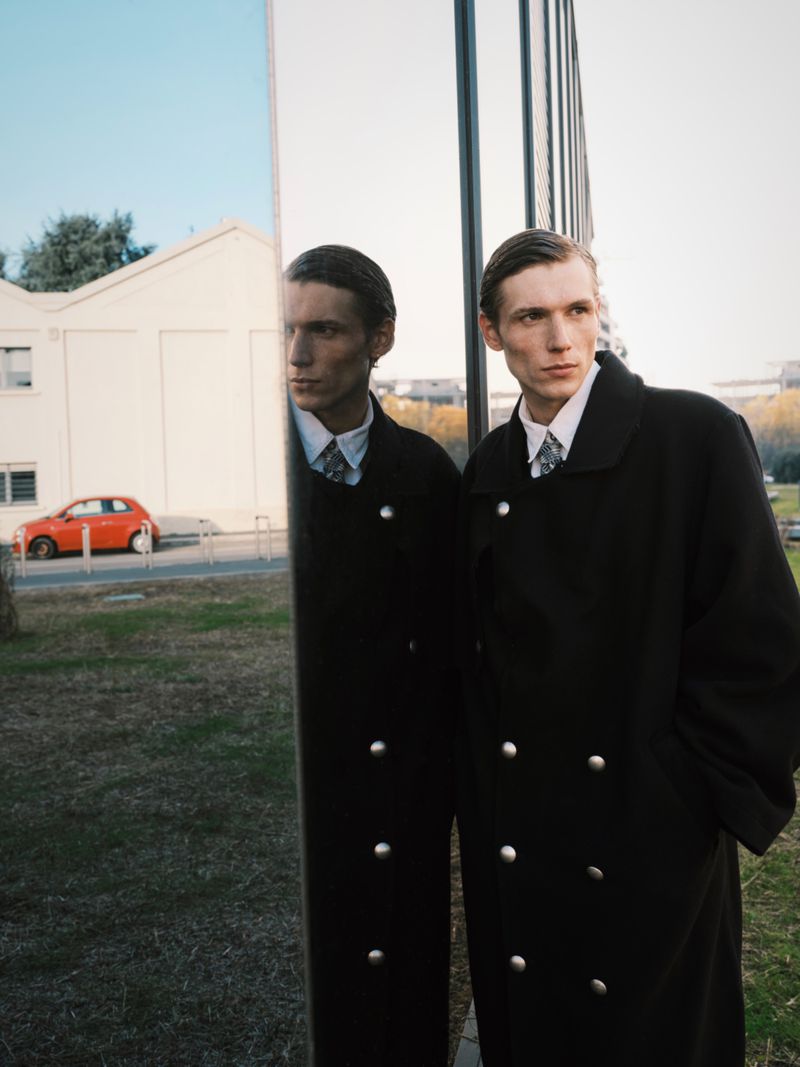 Adult male in long coat and tie stands next to a reflective surface in an outdoor setting with a building and car in background.