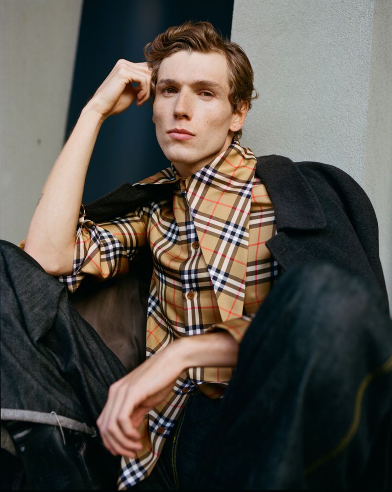 Male model in Burberry plaid shirt and dark jacket poses in outdoor portrait with neutral expression and arm raised to head.