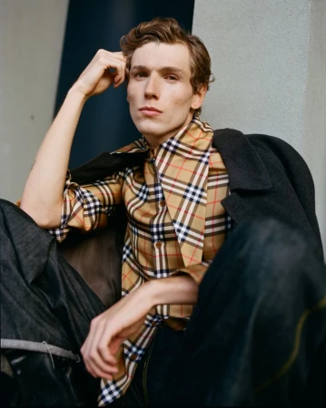 Male model in Burberry plaid shirt and dark jacket poses in outdoor portrait with neutral expression and arm raised to head.