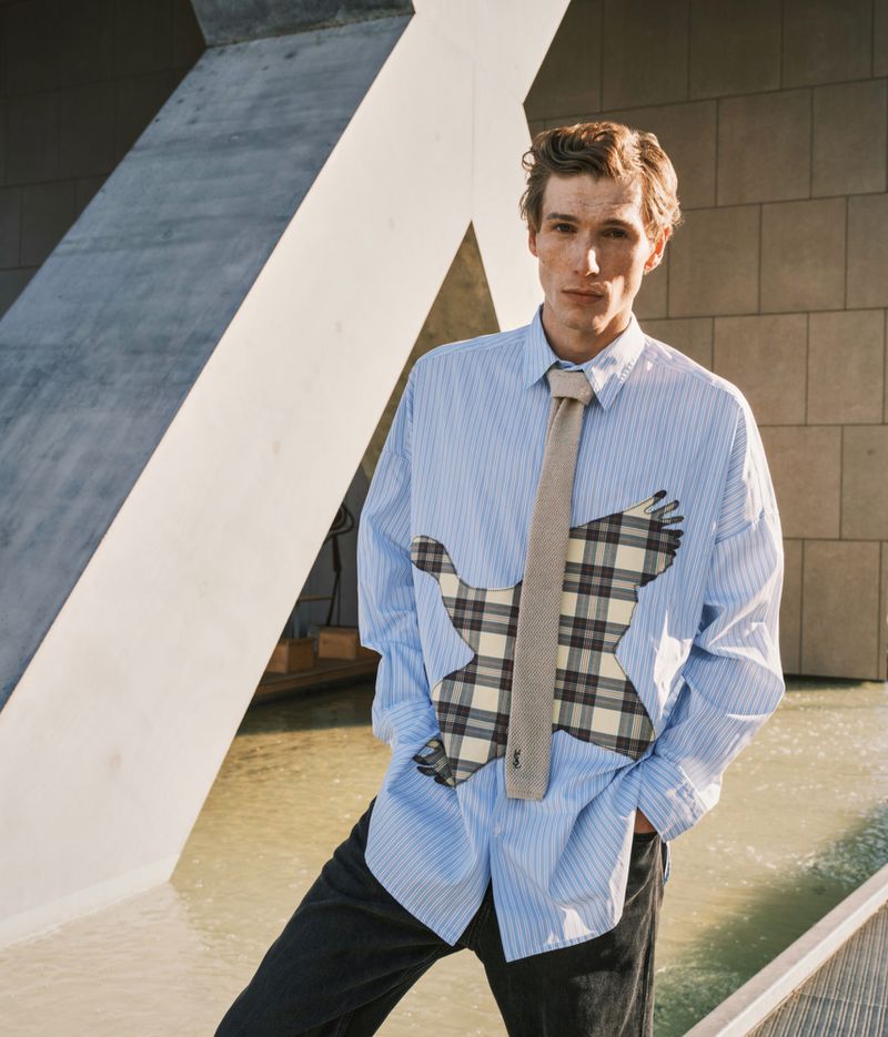 A stylish young adult stands outdoors in a blue striped shirt with a graphic, and tie, against a textured, concrete background, showcasing a casual yet fashionable look.