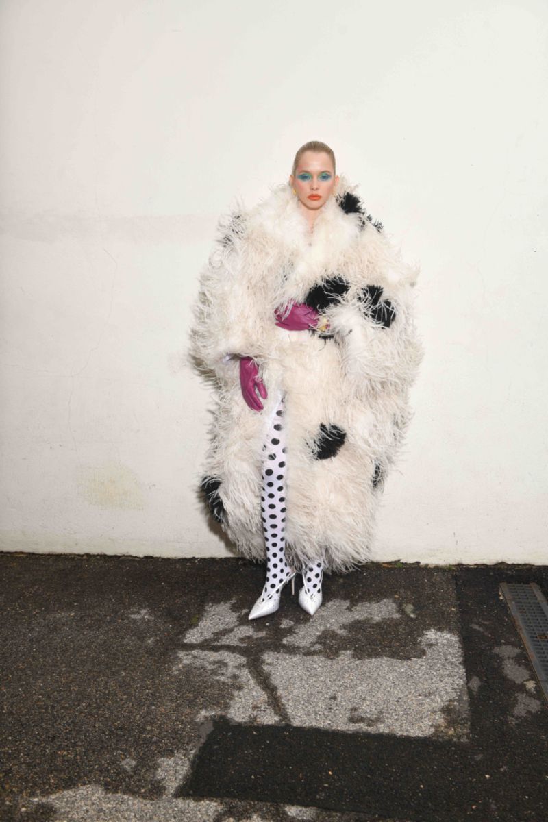 Fashionable woman wearing a large white feather coat with black spots, patterned tights, and bright makeup poses against a white wall.