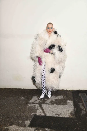 Fashionable woman wearing a large white feather coat with black spots, patterned tights, and bright makeup poses against a white wall.
