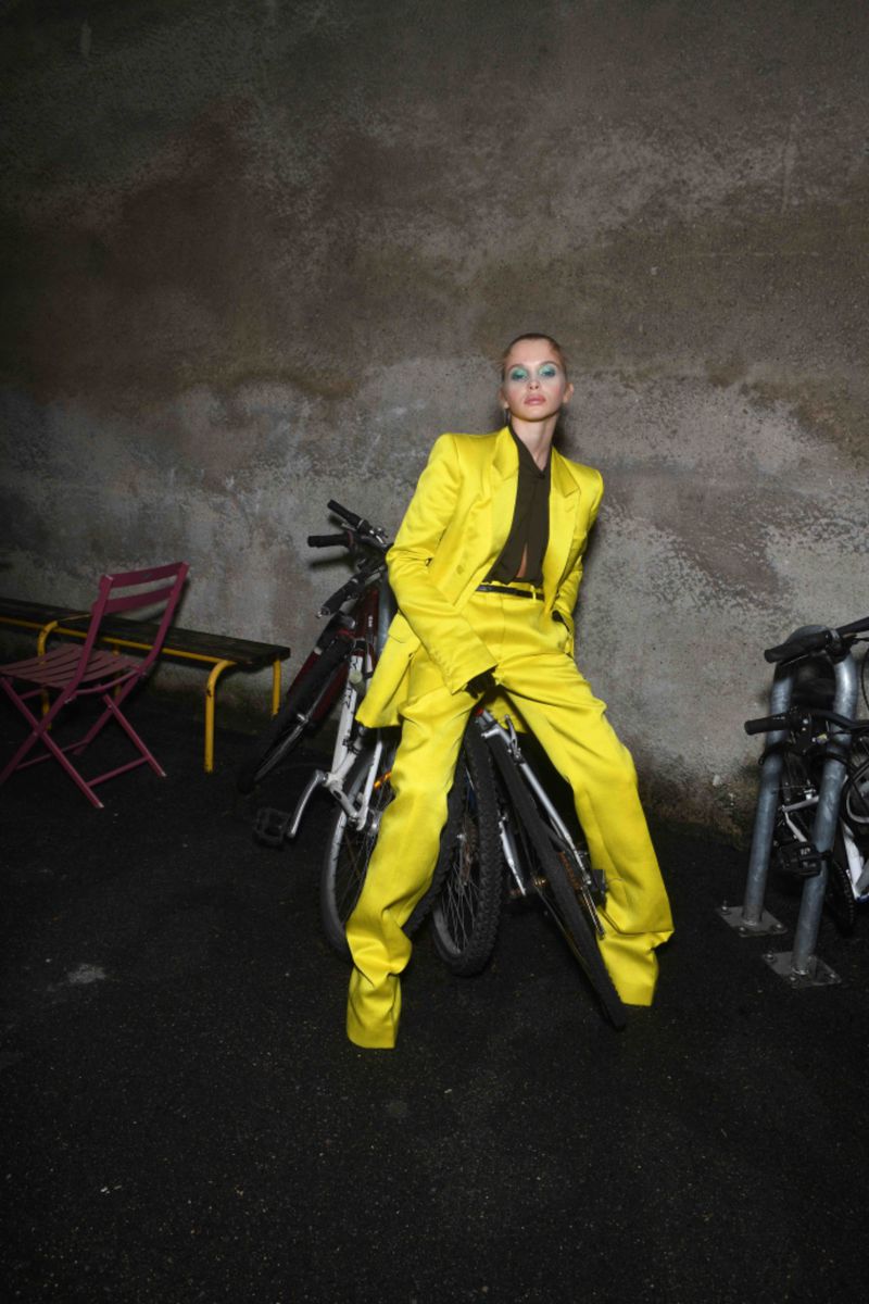 Woman in a yellow suit poses on a bicycle in an urban setting with a weathered wall in the background at night.