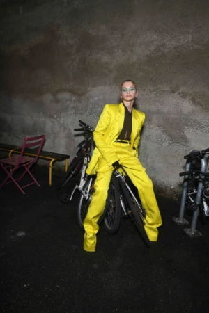 Woman in a yellow suit poses on a bicycle in an urban setting with a weathered wall in the background at night.