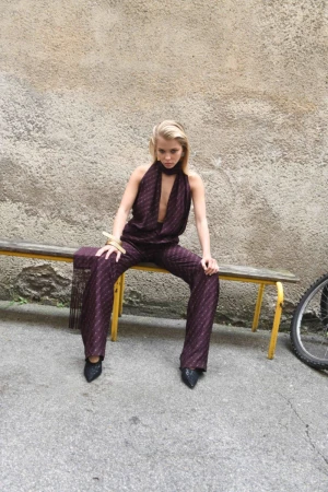 A slender blonde woman sits on a bench in a patterned jumpsuit, paired with high heels and gold bracelet, against a concrete background.