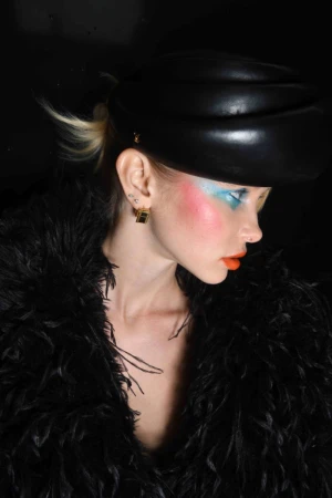 Profile view of a woman in a black hat and feathery coat with colorful makeup in a studio with a dark background and dramatic lighting.
