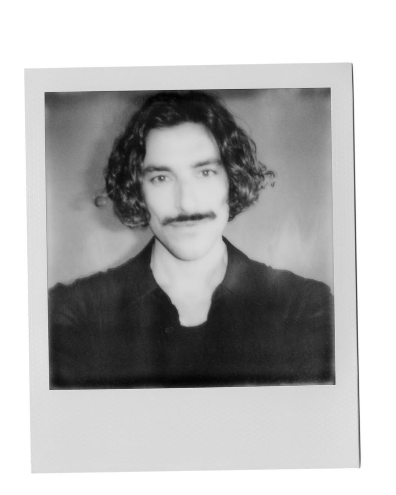 Monochrome portrait of an adult male with a mustache and curly hair, framed in a classic Polaroid-style photo against a neutral backdrop.
