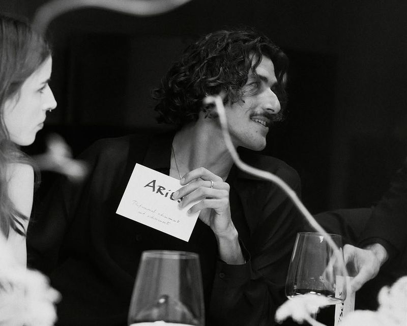 Black and white photograph of a man holding a card with text while sitting at a table with wine glasses and a woman nearby.