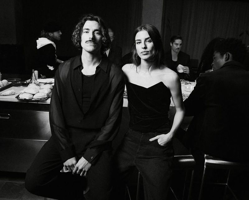 Black and white portrait of a fashionable couple indoors, with other people at tables in the background, creating a social scene.