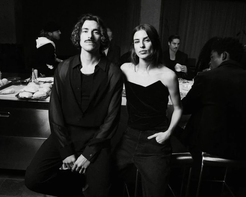 Black and white portrait of a fashionable couple indoors, with other people at tables in the background, creating a social scene.