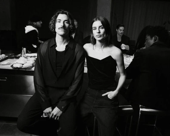 Black and white portrait of a fashionable couple indoors, with other people at tables in the background, creating a social scene.