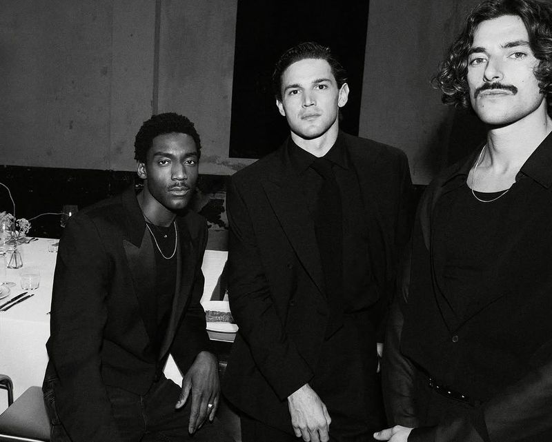 Three stylish men in black formalwear pose indoors, showcasing fashion and portrait photography with a focus on sharp details and monochrome tones.