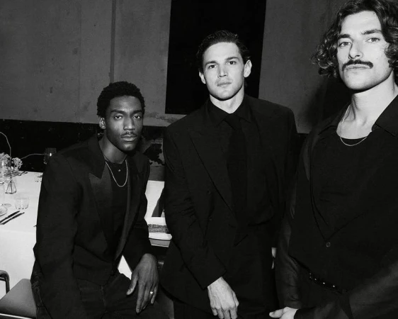 Three stylish men in black formalwear pose indoors, showcasing fashion and portrait photography with a focus on sharp details and monochrome tones.