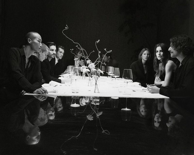 A monochromatic dinner party photo showing a diverse group of adults seated at a white tablecloth-covered table set with glassware.