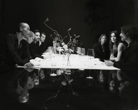 A monochromatic dinner party photo showing a diverse group of adults seated at a white tablecloth-covered table set with glassware.