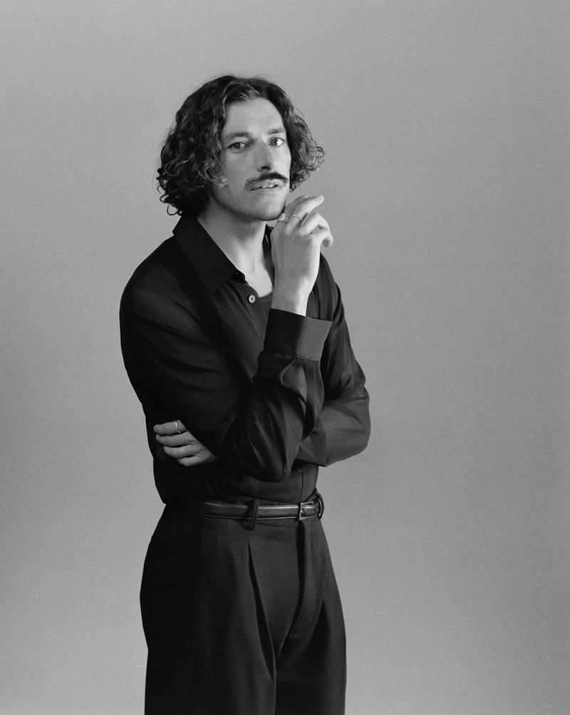 Monochrome portrait of a man with curly hair and a mustache wearing a black shirt against a plain grey backdrop.