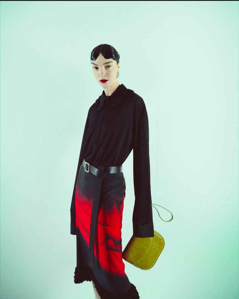 Fashionable woman in black blouse, belted black and red skirt, and olive-green purse stands in a photo studio with a light-blue background.