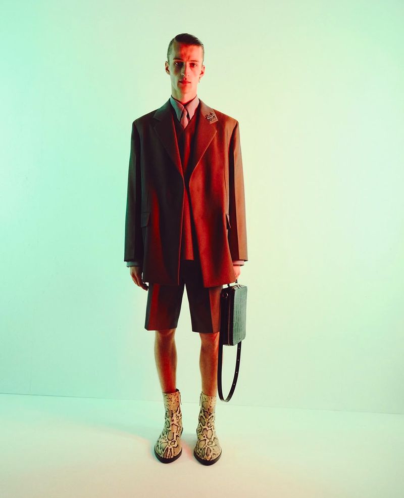 Young man stands in a studio wearing a blazer, shirt, shorts, and snakeskin print boots, holding a black briefcase against a two-tone backdrop.