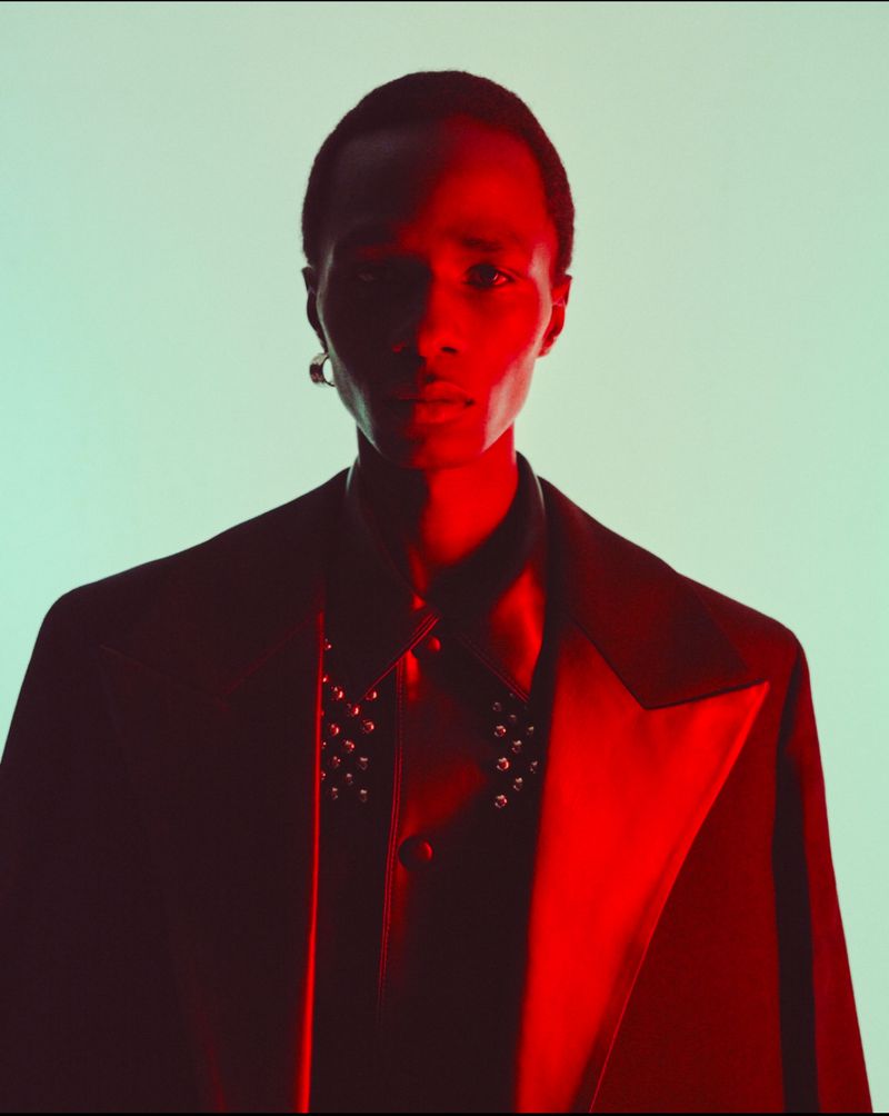 Close-up portrait of a black male model in a dark jacket and shirt with silver studs, lit with contrasting red and green studio lights.