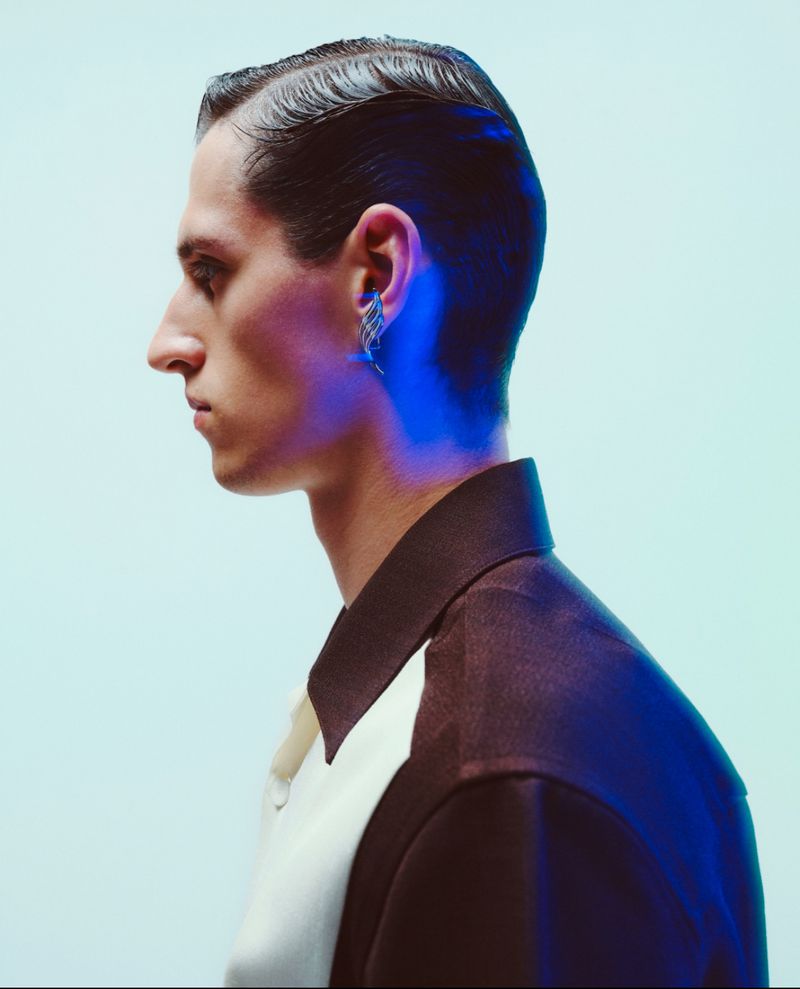 Profile portrait of a man with slicked-back dark hair, wearing an earring and a two-tone collared shirt against a light background.