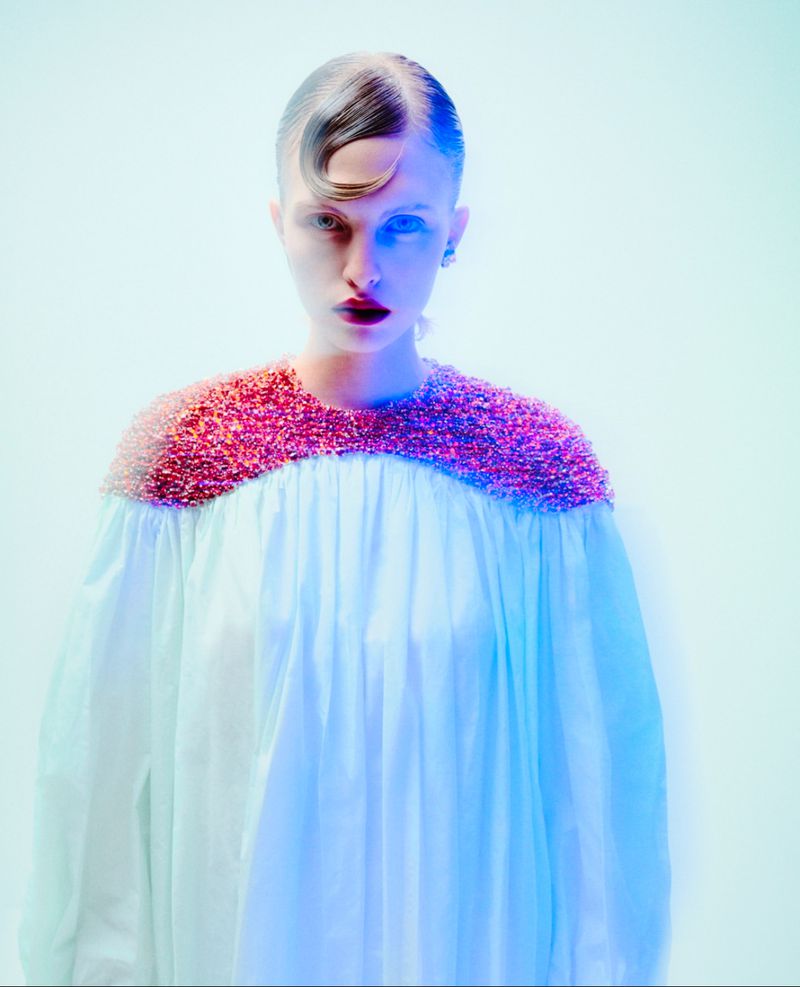 Close-up of a fashion model wearing an embellished white gown with pink trim, against a vivid blue and white backdrop in a studio setting.