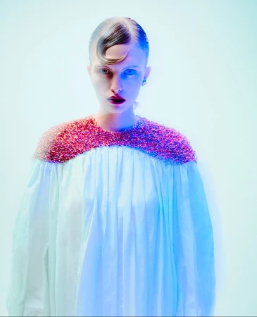 Close-up of a fashion model wearing an embellished white gown with pink trim, against a vivid blue and white backdrop in a studio setting.