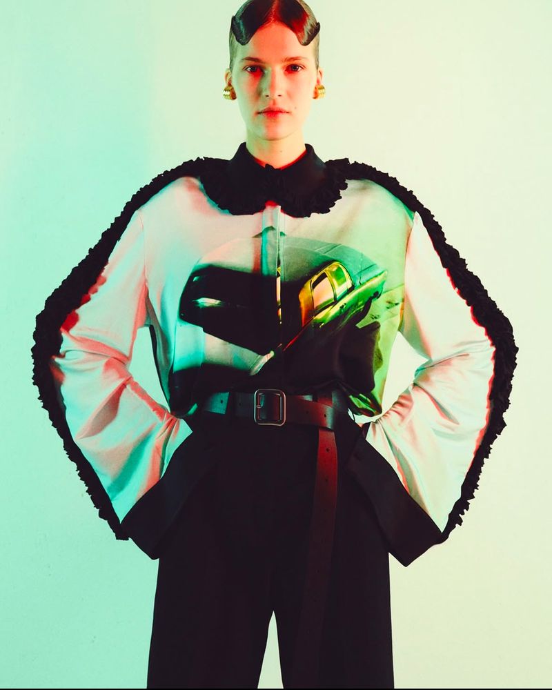 Woman posing in a studio wearing a printed blouse with ruffled trim, black pants and a belt with gold earrings against a green backdrop.