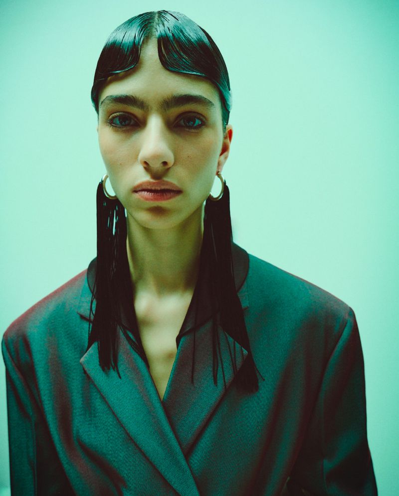 Fashion portrait of a woman with short, wet-look black hair, statement earrings, and a teal suit jacket against a teal backdrop.