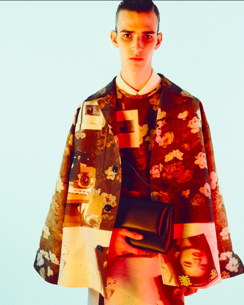 Male model wears a floral print coat and a black bag. He is set against a light blue backdrop in a fashion studio shot.