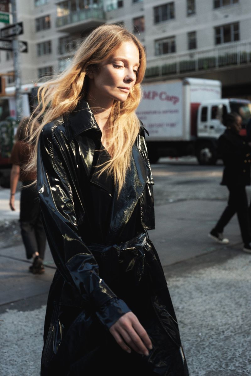 Eniko Mihalik walking down a New York street wearing a glossy black trench coat in a fashion editorial photograph by Adam Jelen.