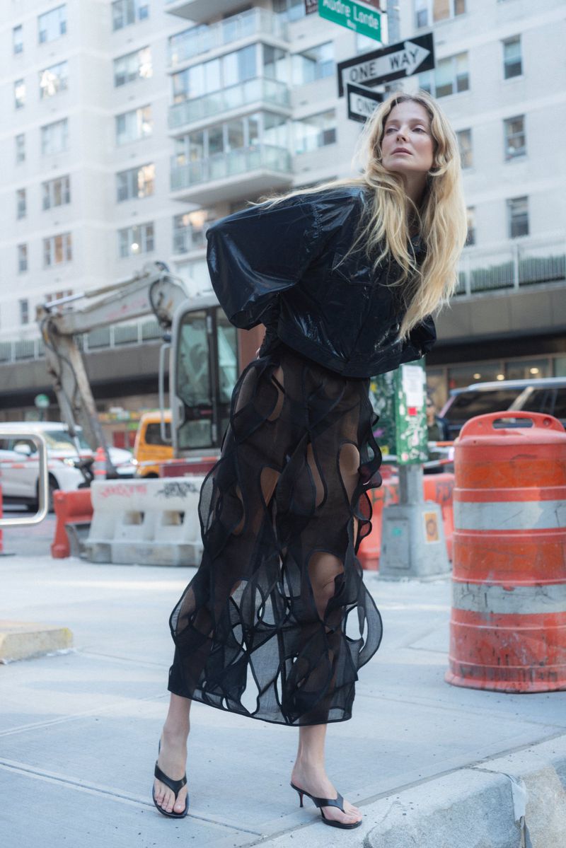 Adult female fashion model with long blonde hair poses in a shiny black top and semi-sheer black skirt on a city sidewalk near construction.