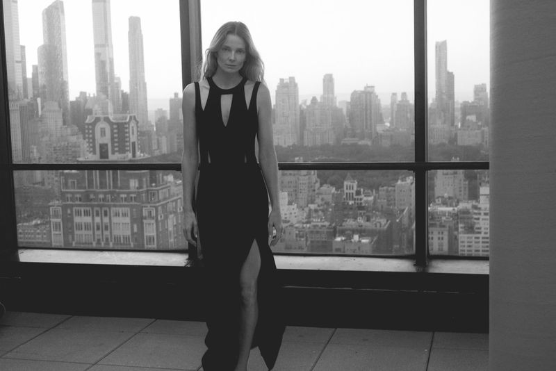 Eniko Mihalik wearing a black cutout dress standing before a New York City skyline window