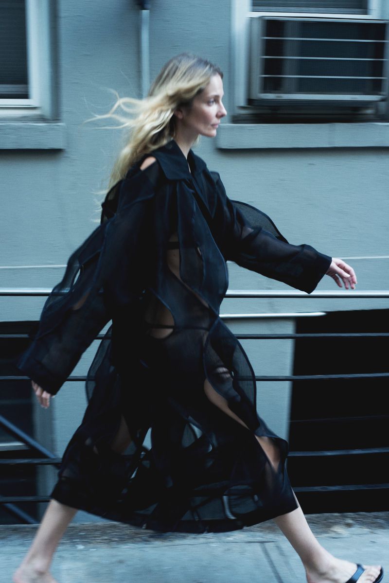 A slender woman with blonde hair walks along a city street wearing a long, sheer black coat-dress, photographed with motion blur.