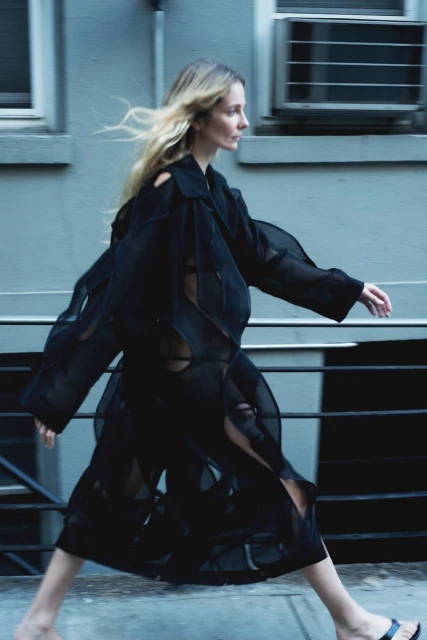 A slender woman with blonde hair walks along a city street wearing a long, sheer black coat-dress, photographed with motion blur.