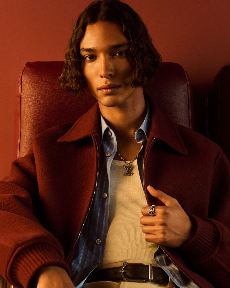 Portrait of a dark-haired male model with a burgundy jacket sitting in a red leather chair in front of a red background.