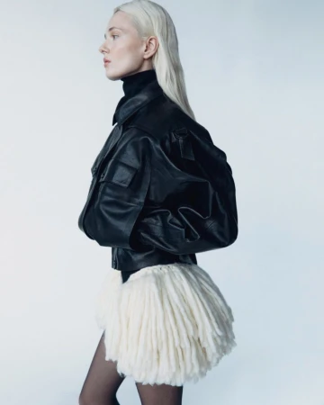 Fashion shot of a slender woman with light blonde hair wearing a black jacket and short white skirt against a bright neutral backdrop.