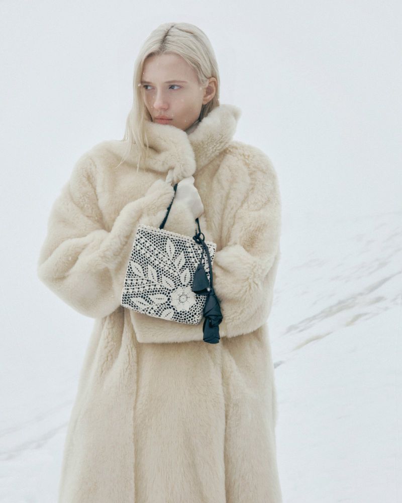 A fair-skinned woman with blonde hair wears a cream-colored faux fur coat and holds a beaded purse with tassels in a snowy outdoor scene.