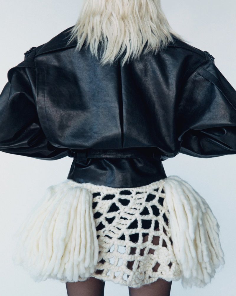 Back view of a fashion model wearing a black leather jacket and a short, cream-colored, crocheted skirt against a white background.