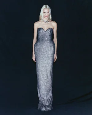 Model with platinum blonde hair standing in a strapless floor-length metallic silver gown against a dark background.