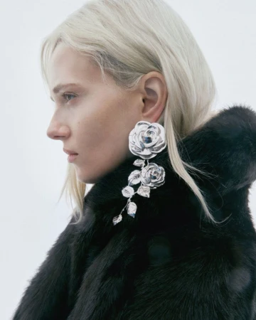 Profile portrait of a blonde woman wearing a large sculptural silver rose earring and a black textured fur coat.
