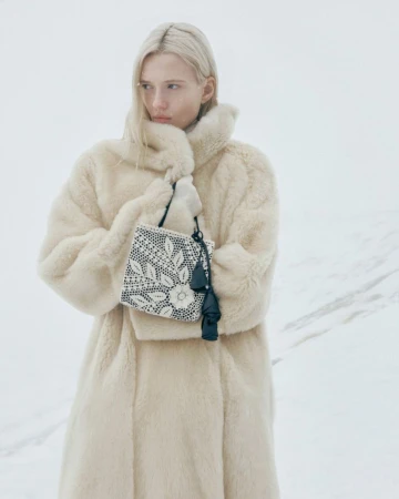 Blonde model in an oversized cream fur coat holding a black and white floral handbag against a snowy landscape.