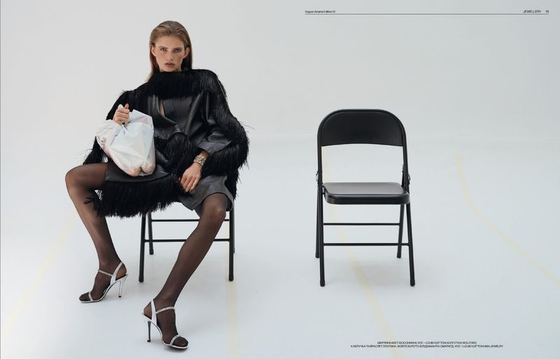 Fashion editorial shot of a woman in a black fringed coat and heels sitting on a chair holding a plastic bag in a white studio.