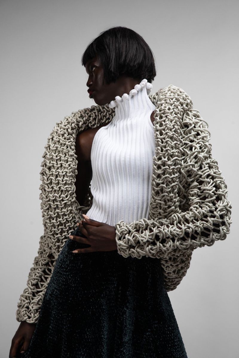 Fashion shot: A black woman poses in a white turtleneck, and a large crocheted jacket against a neutral grey background in a studio setting.